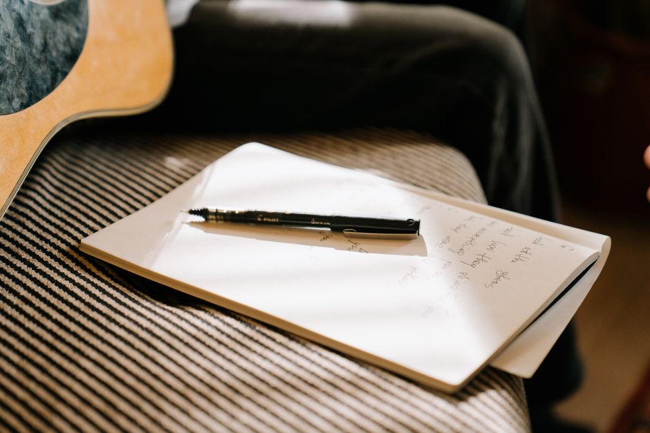 A close-up of a guitar, notebook, and pen suggests a moment of creative songwriting indoors.